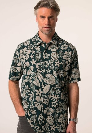 Uomo che indossa una camicia verde a maniche corte con motivi floreali tropicali beige, con le mani parzialmente nelle tasche, su sfondo semplice.