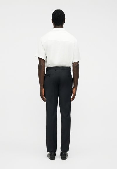 Homme debout de dos, portant une chemise blanche à manches courtes, un pantalon de costume noir avec des poches arrière, et des chaussures de ville noires, sur un fond uni.