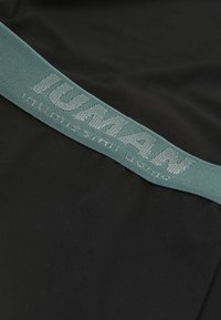 Una tela negra con una textura suave presenta una cintura elástica en color azul verdoso que muestra "HUMAN" en bordado gris claro, diseñada para comodidad y ajuste.