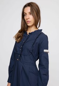 Hosszú barna hajú nő, tengerészkék ruhában, fodrokkal, gombokkal és csipkedett részlettel a felső karján, oldalra néz.