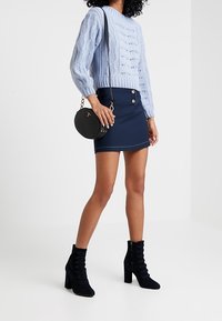 Světle modrý svetr s pleteným vzorem, námořnická sukně na knoflíky, černá kulatá kabelka a černé semišové kotníkové boty s nabraným detailem.