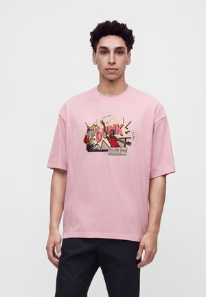 Junger Mann mit kurzen lockigen Haaren, der ein pinkes T-Shirt mit einem Comic-Stil Basketball-Dunk-Grafik und schwarze Hose trägt.
