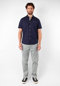 Chemise bleue marine à manches courtes avec une poche sur la poitrine, associée à un pantalon gris clair et des baskets noires avec des semelles blanches.