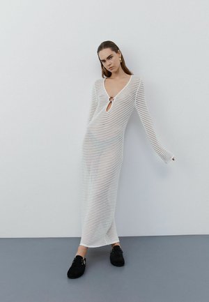 Kobieta w długiej białej dzianinowej sukience z głębokim dekoltem w serek i detalem w formie otworu, stojąca przy gładkiej białej ścianie i szarej podłodze.