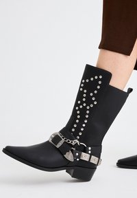 Botas hasta la rodilla de color negro, hechas de un material texturizado, con tachuelas plateadas dispuestas en un patrón decorativo y un acento de cadena metálica con una hebilla.