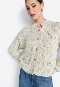 Helle cremefarbene Strickjacke mit mehrfarbigen Sprenkeln in Blau und Gelb, silbernen Knopfverschlüssen und zwei Taschen vorne. Lange Ärmel.