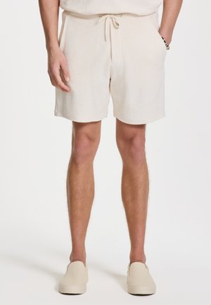 Beige fleece shorts with an elastic waistband and drawstring. Side pockets and a relaxed fit. Paired with light slip-on shoes.