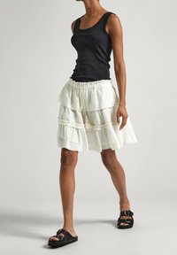 Schwarzes, geripptes Trägershirt und einen geschichteten weißen Baumwollrock mit Rüschendetails. Schwarze Sandalen runden das Outfit ab. Neutraler Hintergrund.