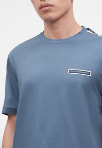 Άνδρας που φορά μπλε μπλούζα EA7 Emporio Armani με λογότυπο στο στήθος και ρίγα στον ώμο, κοντινό πλάνο στο άνω σώμα.