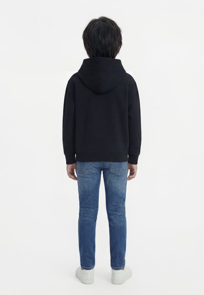 Παιδί με την πλάτη γυρισμένη, φοράει μαύρη φούτερ με κουκούλα, μπλε τζιν και λευκά αθλητικά παπούτσια, με λευκό απλό φόντο.