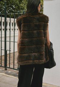 Vesta din blană maro cu o textură cu benzi, design fără mâneci și un aspect moale. Asortată cu o geantă neagră țesută și pantaloni negri largi.