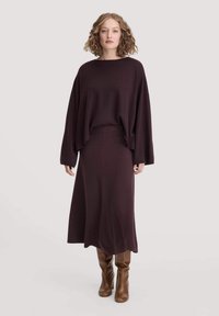Burgundy neulepusero, jossa on leveät hihat, yhdistettynä matching pitkään hameeseen. Mallilla on jalassaan ruskeat polven yli ulottuvat saappaat. Pehmeä rakenne ja virtaava muotoilu.