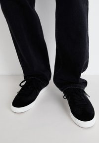 Schwarze Wildledersneaker mit strukturierten Einsätzen, weißen Gummisohlen und flachen Schnürsenkeln, kombiniert mit locker sitzenden schwarzen Jeanshosen.
