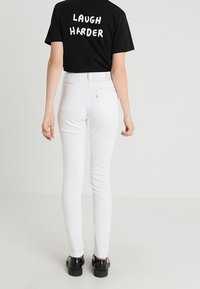 Camiseta negra con el texto "RIÈTE MÁS FUERTE" en blanco, combinada con unos jeans ajustados blancos de cintura alta. Modelo lleva zapatos de plataforma negros.