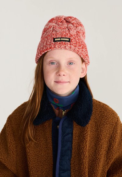 Jeune personne avec des cheveux roux portant un bonnet tricoté rouge et blanc avec une étiquette, un manteau marron texturé et un col roulé bleu sur un fond uni.
