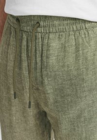Elastična pasica jogger hlač v zeleni herringbone vzoru, ki ima vrvico z temnimi konci in mehak teksturiran material.