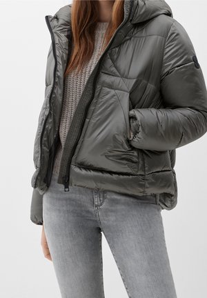 Femme portant une doudoune grise brillante par-dessus un pull beige en tricot et un jean gris clair, main dans la poche de la veste.