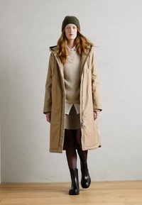 Cappotto imbottito beige con cappuccio, abbinato a un maglione a coste e una gonna. Il modello indossa stivali neri in gomma e un berretto verde scuro.