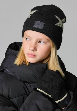 Bonnet en tricot noir avec un motif d'étoiles argentées et une étiquette noire ; gants assortis avec une rayure argentée, portés avec un manteau noir rembourré.