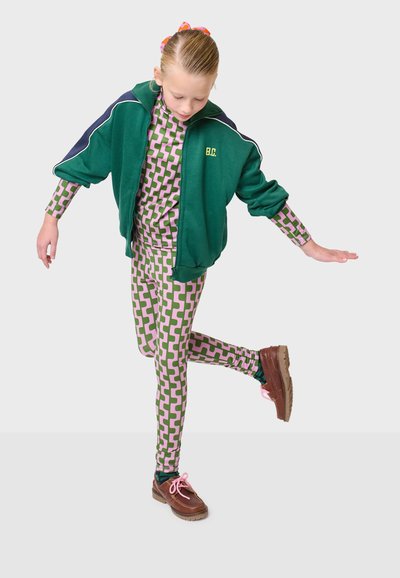 Grüne Zip-Jacke mit dunkelblauen Akzenten, kombiniert mit einem bodenlangen Anzug mit pinkem und grünem Muster und Leggings. Braune Schuhe mit pinken Schnürsenkeln.