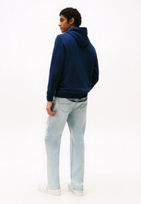 Marineblå hettegenser med snøring, kombinert med lyseblå rette jeans og hvite sneakers, med synlige bakstingdetaljer.