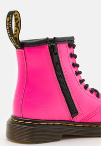 Bota sintética de tobillo en rosa con detalles en negro, que incluye una cremallera lateral, cordones negros, costuras amarillas y una suela negra texturizada.