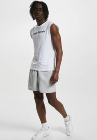 Hvit ermeløs skjorte med svart logo, kombinert med lysegrå shorts og hvite sneakers. Glatt tekstur, uformell atletisk stil.