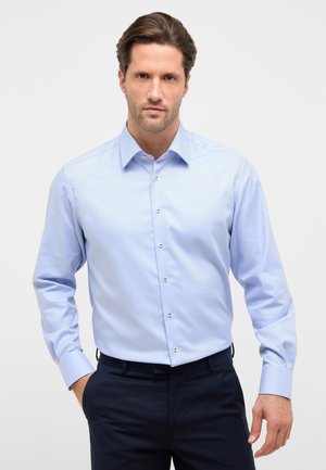 Mand iført lyseblå skjorte med knapper, stukket ned i mørkeblå bukser, står med den ene hånd i lommen mod hvid baggrund.