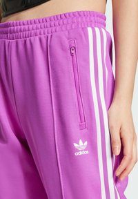 Lila träningsbyxor med texturerad yta, med vita sidränder, en ficka med dragkedja och en liten vit Adidas-logotyp.