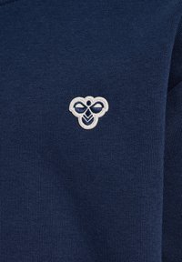 Marinblå stickad tröja med ribbad textur. Har en liten, broderad logotyp i vitt garn som föreställer en symmetrisk design.