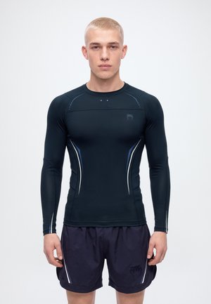 Un jeune homme se tient face à l’objectif, vêtu d’un haut de sport noir à manches longues et d’un short foncé, sur un fond clair uni.