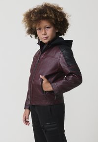 Giacca in faux leather bordeaux con accenti neri, tasche con zip e cappuccio. Presenta una texture liscia e un design a coste sulle spalle.