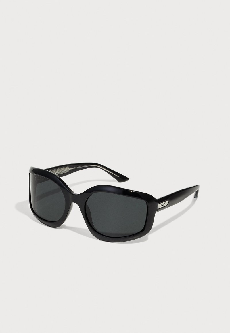 Óculos de sol oversized pretos com lentes escuras, armação de plástico suave e hastes finas. Apresenta um detalhe de logo metálico na lateral.