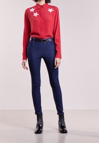 Červená košile s dlouhými rukávy a bílými hvězdnými aplikacemi na hrudi, spárovaná s tmavě modrými přiléhavými džíny a černými kotníkovými botami s žebrovanou texturou.