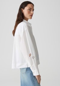 Witte blouse met lange, losse mouwen met uitsnijdingen in bladerenpatronen bij de manchetten. De stof lijkt soepel en lichtgewicht, met een kraag.
