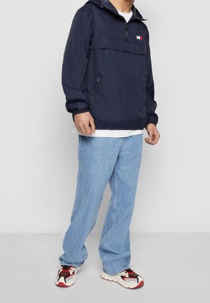 Rompevientos azul marino con capucha, diseño de medio cierre, logo en el pecho y puños elásticos, combinado con vaqueros azul claro y zapatillas multicolores.