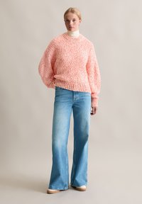 Rosa und weiß gestrickter Pullover mit strukturiertem Muster und weiten Ärmeln, kombiniert mit hellblauen Schlagjeans und beigen Sneakers. Glattes, lässiges Erscheinungsbild.