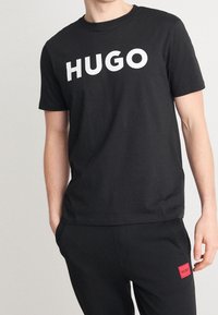 Camiseta de algodón negra con un gran logo blanco "HUGO" en el pecho, cuello redondo y mangas cortas. Llevada con pantalones negros.