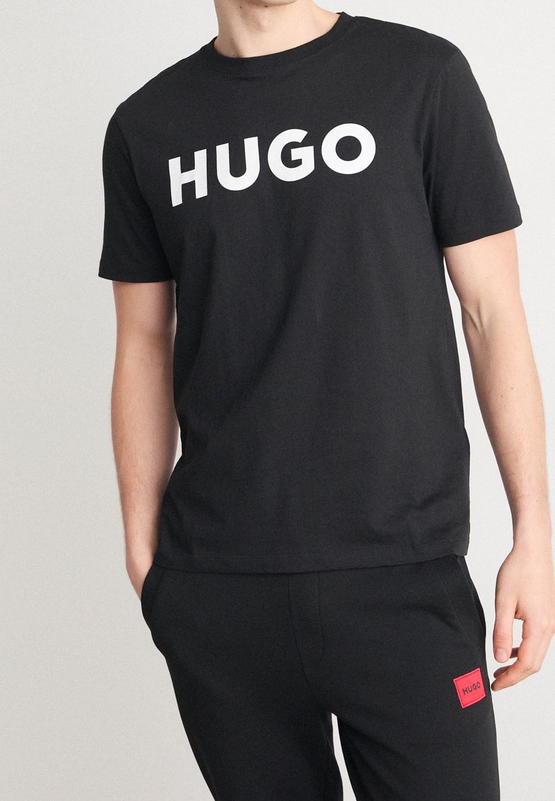Camiseta de algodón negra con un gran logo blanco "HUGO" en el pecho, cuello redondo y mangas cortas. Llevada con pantalones negros.