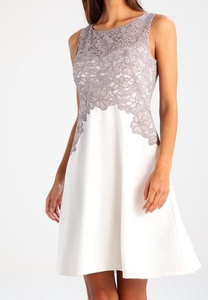 Biała sukienka bez rękawów z rozkloszowaną spódnicą długości do kolan i lawendową kwiecistą koronkową nakładką na górnej części. Model ukazany od szyi do kolan.