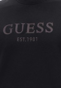 Μαύρο ύφασμα με ανάγλυφη κεντητή επιγραφή "GUESS" πάνω από το "EST. 1981" σε σκούρες γκρι κλωστές.