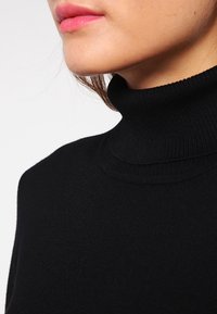 Pull noir à col roulé avec une texture côtelée, un col haut et une coupe ajustée. Gros plan montrant les détails du col et le tissu lisse.