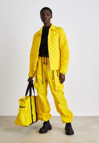 Casaco amarelo acolchoado e calças cargo combinando, ambas com cordões ajustáveis, acompanhados de uma bolsa tote amarela com detalhes em preto.