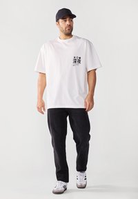 Camiseta blanca de algodón con estampado gráfico negro en el pecho; combinada con pantalones de mezclilla oscuros y zapatillas blancas con detalles y logotipo en negro.