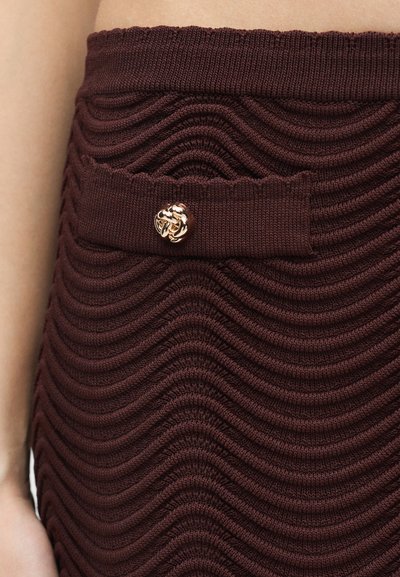 Gros plan sur une jupe en tricot bordeaux côtelé avec des motifs ondulés et une boucle décorative dorée sur un rabat en tissu à la taille.