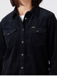Black corduroy shirt with a snap button front, two chest pockets, and a subtle logo. Features ribbed texture and pointed collar design.