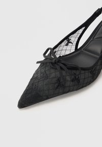Escarpins slingback en filet noir avec un bout pointu, présentant un motif en grille et une broderie florale. Accent nœud noir sur le devant.