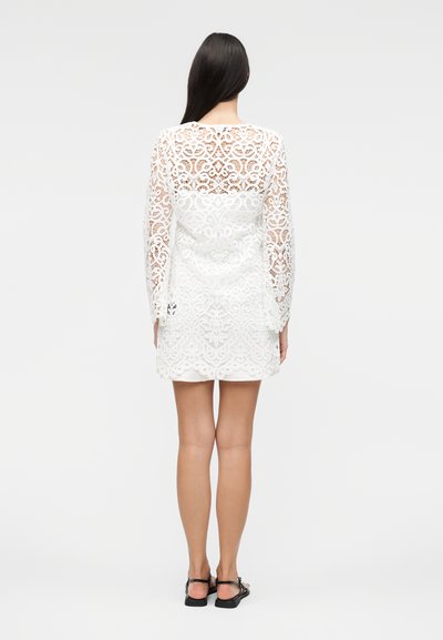 Femme aux longs cheveux foncés portant une robe blanche en dentelle à manches longues et des sandales noires, debout, dos à un fond uni.