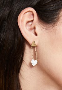 Guld hjärtformade stiftörhängen med en hängande kedja som har en klar kristallhjärtcharm. Släta metall- och glittrande texturdetaier.