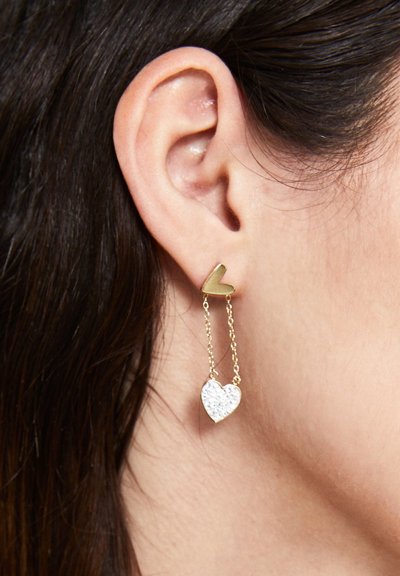 Guld hjärtformade stiftörhängen med en hängande kedja som har en klar kristallhjärtcharm. Släta metall- och glittrande texturdetaier.